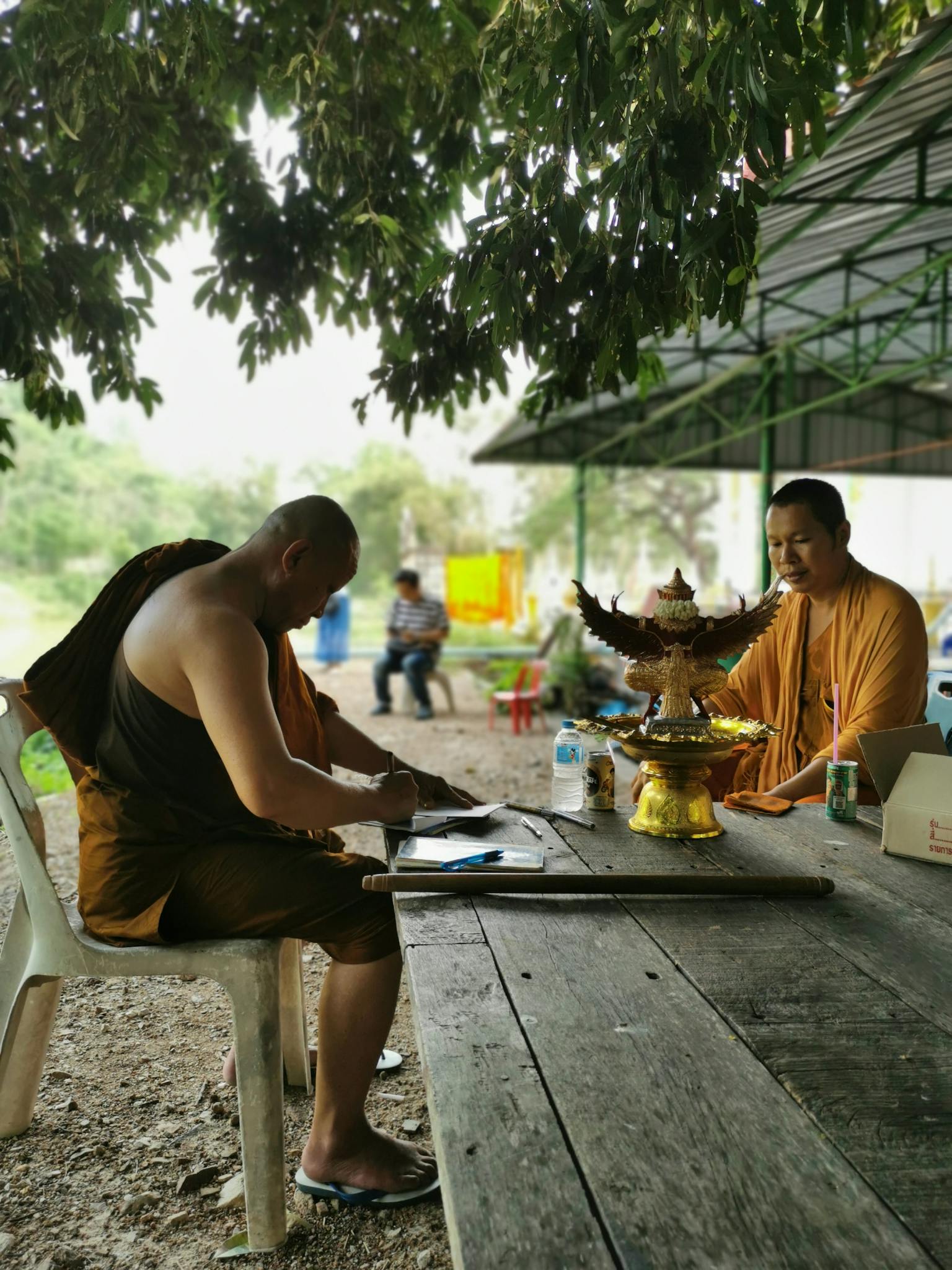 Two monks sitting at a table under a tree in Tambon Wang Lum, Thailand, embracing tranquility.
